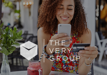 Very Group Thumbnail with logo. Women looking at a smartphone to make a payment.